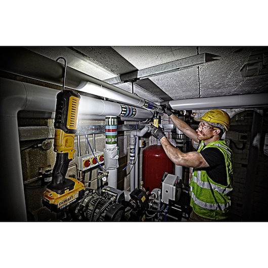 A worker wearing a safety vest and helmet is installing or repairing pipes in an industrial setting. A DEWALT DCL050 portable work light is illuminating the workspace.