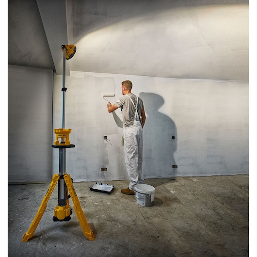 A painter uses a paint roller on a wall, illuminated by a yellow DEWALT tripod work light in a room under renovation.
