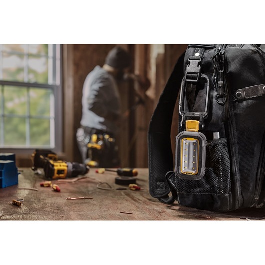 A DEWALT portable LED work light (model DCL182-KR) clipped to the side of a black tool bag on a wooden workbench, with various tools and electrical parts scattered on the table. A person is working in the background near a window.