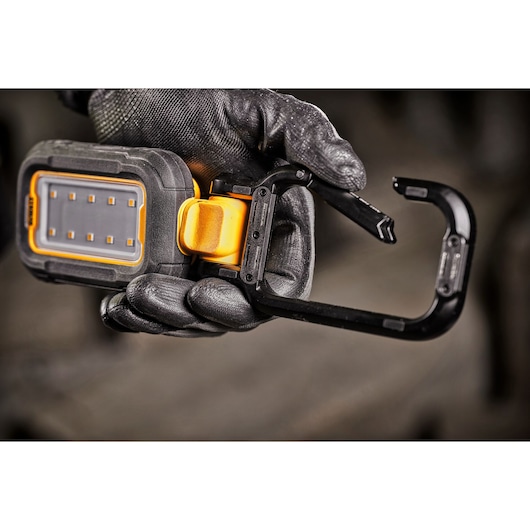 A gloved hand holding a DEWALT portable LED work light with a large black carabiner clip attached.