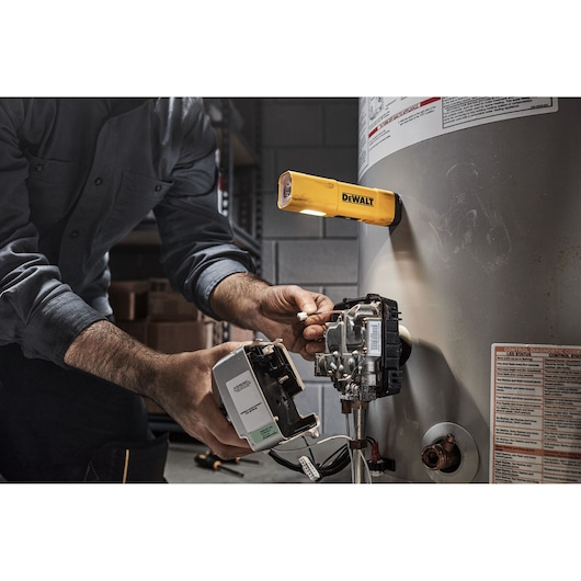 A person is repairing a water heater with a yellow DEWALT work light attached to the heater, illuminating the workspace.