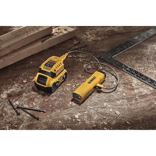 DEWALT DCL183-KR flexible LED work light with a detachable battery pack connected by a cable, placed on a wooden workbench with wood shavings, nails, and measurement tools.