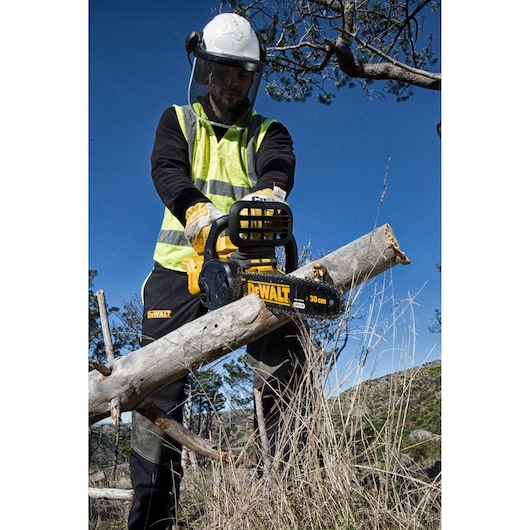 A person wearing protective gear and high-visibility clothing is using a DEWALT DCM565N chainsaw to cut a tree branch outdoors. The chainsaw has a visible 30 cm blade and is held securely, with the surroundings showing grass and trees.