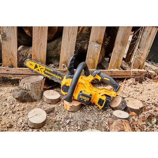 A yellow DEWALT XR brushless cordless chainsaw is placed on the ground surrounded by cut wooden logs and wood shavings. The chainsaw is fitted with an 18V battery and is resting outdoors in front of a wooden fence.