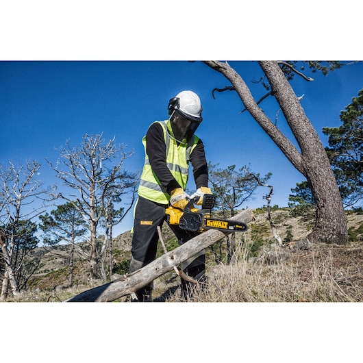 A person wearing safety gear and a high-visibility vest is using a DEWALT DCM565N chainsaw to cut a fallen tree branch outdoors in a forested area.