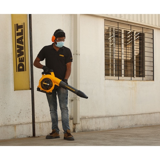 A person wearing hearing protection and a DEWALT-branded shirt is standing outdoors next to a DEWALT sign, holding a yellow DEWALT DCM572N cordless leaf blower. The blower is pointed towards the ground near a building with barred windows.