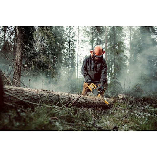 Person using a DEWALT chainsaw to cut a fallen tree in a misty forest, wearing safety gear.