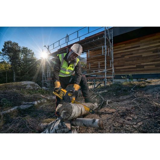 Worker uses a DEWALT chainsaw to cut logs outdoors, wearing safety gear near a wooden building and scaffold.