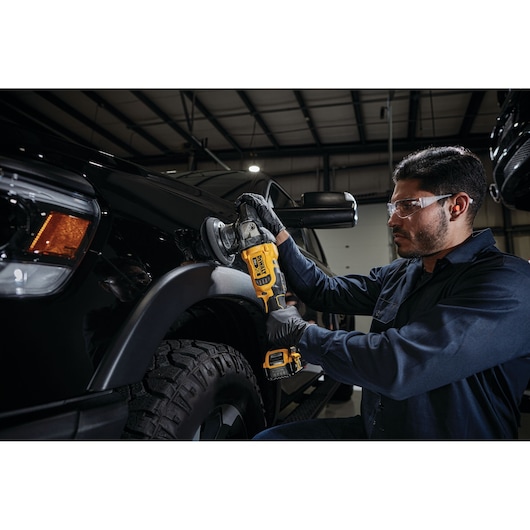 A person uses a DEWALT cordless polisher (model DCM848P2) to polish the fender of a black vehicle in an indoor garage. The person is wearing gloves and a dark shirt, and their face is blurred.