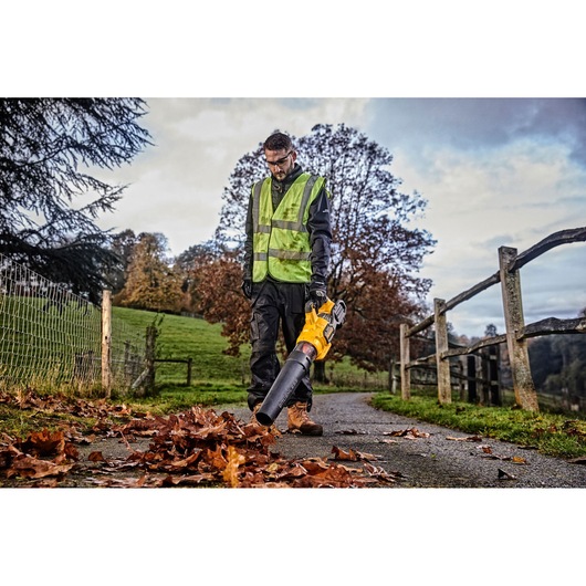 A person wearing a high-visibility vest uses a yellow DEWALT leaf blower (model DCMBA572X1) to clear fallen leaves on an outdoor path, with greenery and trees in the background.