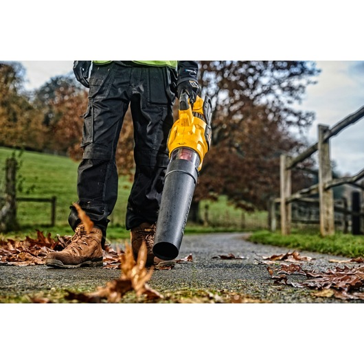 A person wearing work boots and gloves is using a yellow DEWALT leaf blower to clear leaves from a pathway outdoors. The scene includes autumn foliage, a wooden fence, and grassy areas.