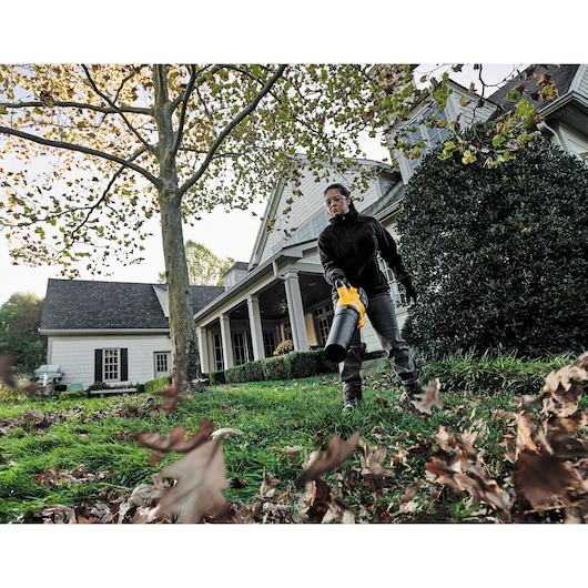 A person is using a DEWALT leaf blower to clear fallen leaves from a lawn in front of a house. The individual is standing outdoors near a large tree, wearing a dark jacket and gloves. The house has white walls and a porch with columns.