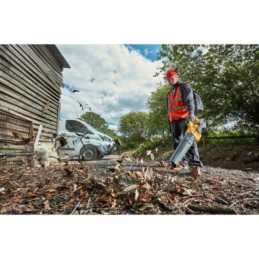 A person wearing a red safety vest and helmet is using a DEWALT leaf blower (model DCMBA572X1) to clear leaves and debris from the ground near a wooden structure and a white van, outdoors under a cloudy sky.