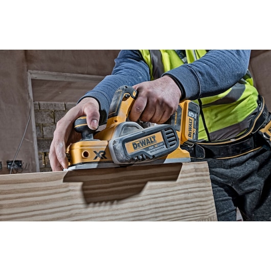 Worker uses a DEWALT cordless planer on a wooden plank in a construction setting.