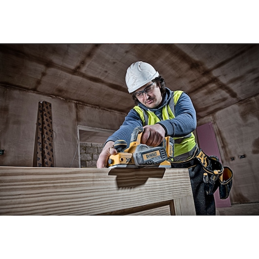Worker uses DEWALT DCP580N planer on wood indoors, wearing safety gear and tool belt.