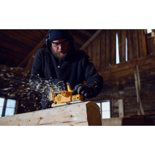 A person wearing protective gloves and earmuffs uses a yellow DEWALT DCP580P2 cordless planer to smooth a wooden beam indoors. Wood shavings are flying as the tool operates.