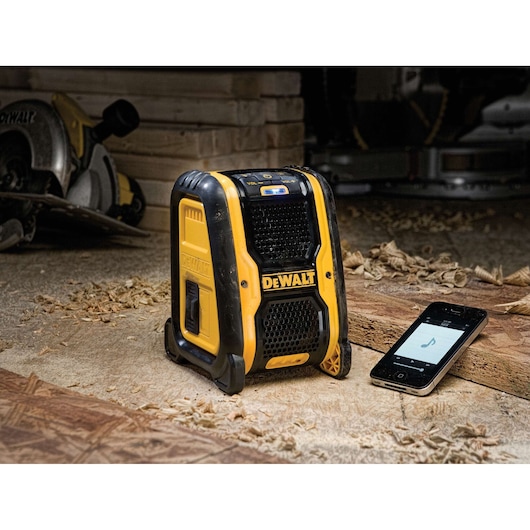 A DEWALT portable Bluetooth speaker (model DCR006) is shown on a woodworking site surrounded by wood shavings, with a smartphone next to it displaying a music app. In the background, there are power tools and stacked wood pieces.