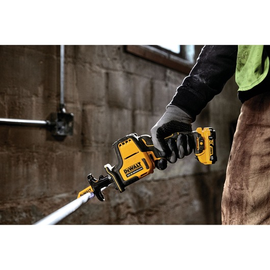 Person using a DEWALT DCS312N cordless reciprocating saw to cut a pipe in a workshop.