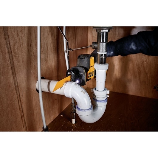 DEWALT tool cutting white PVC plumbing pipe under a sink in a wooden cabinet.
