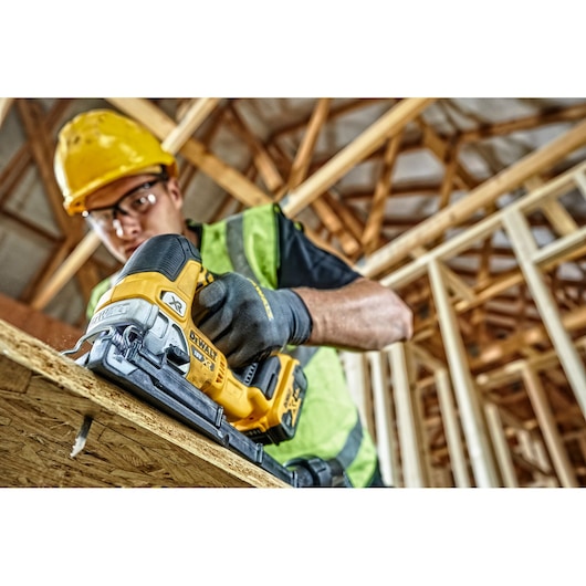 A construction worker wearing a yellow hard hat and safety vest uses a DEWALT DCS334N cordless jigsaw to cut wood at a building site with exposed wooden beams.