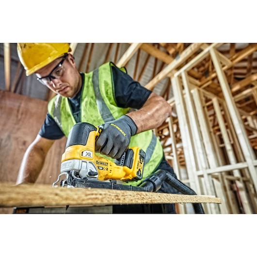 A worker wearing a safety helmet and high-visibility vest uses a DEWALT DCS334N cordless jigsaw to cut a wooden board at a construction site with wooden framing in the background.