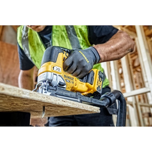 A person wearing work gloves and a safety vest is using a DEWALT DCS334N cordless jigsaw to cut a wooden board at a construction site.