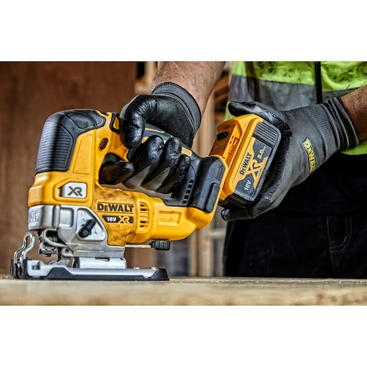 A gloved hand holding a DEWALT 18V XR jigsaw, with a 5.0 Ah battery attached, positioned on a wooden surface in a workshop setting.