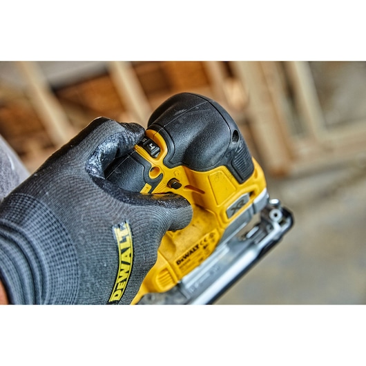 Close-up of a gloved hand adjusting settings on a yellow and black DEWALT power tool, likely a jigsaw, with a blurred workshop background.