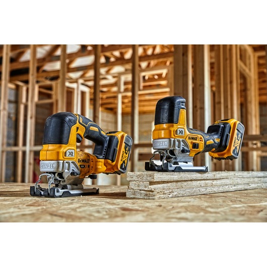 Two DEWALT cordless jigsaws are placed on a wooden surface in a construction site with unfinished wooden framing in the background.