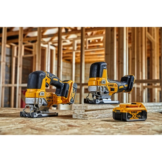 Two DEWALT cordless jigsaws and a DEWALT 18V battery are placed on a wooden surface inside a building under construction with exposed wooden framing.