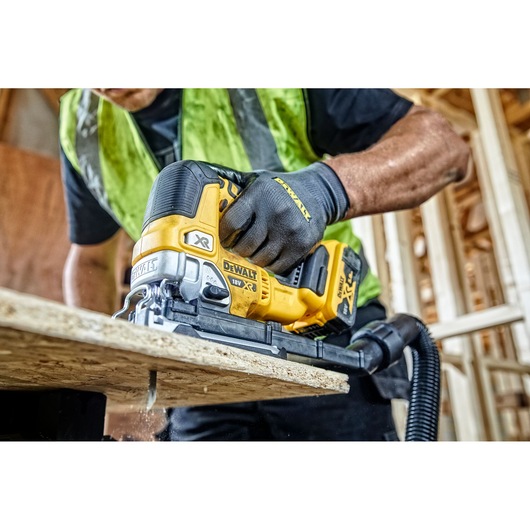A person wearing work gloves and a safety vest uses a DEWALT 18V XR cordless jigsaw to cut through a sheet of wood in a construction setting.