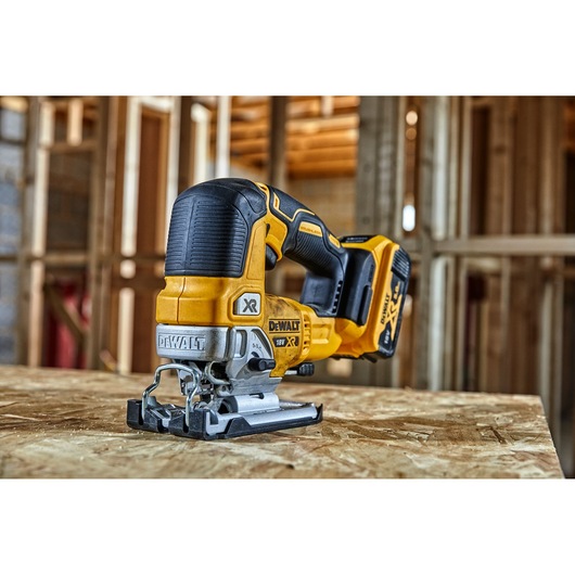 A DEWALT cordless jigsaw model DCS334P2 is placed on a wooden surface in a construction setting. The tool is yellow and black with visible DEWALT branding and an 18V XR lithium-ion battery.