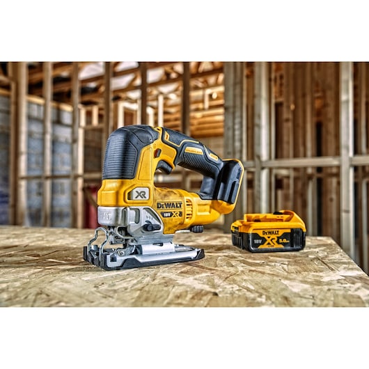 A yellow and black DEWALT cordless jigsaw and a detachable battery are placed on a wooden surface in front of an unfinished construction site framework.