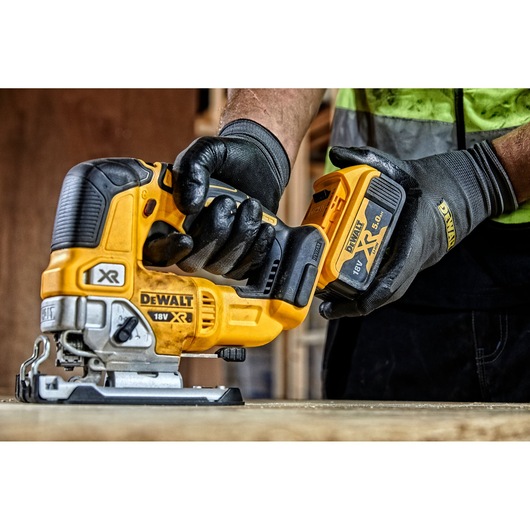 A person wearing black gloves is installing a battery into a yellow DEWALT 18V XR cordless jigsaw, model DCS334P2, on a wooden surface.