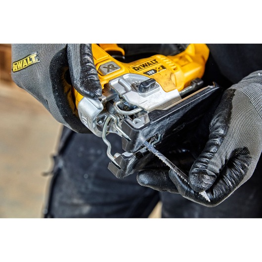 Close-up of a person wearing gloves handling a DEWALT 18V XR jigsaw, preparing to insert a jigsaw blade into the saw.