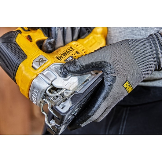 Close-up of a person wearing protective gloves holding a yellow DEWALT cordless jigsaw, model XR 18V, during use.