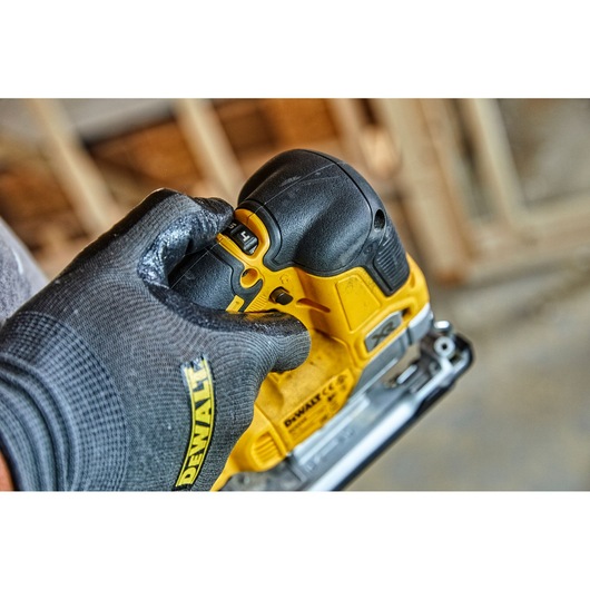 A close-up of a gloved hand adjusting the control on a yellow DEWALT cordless jigsaw, model DCS334P2, with a blurred workshop background.