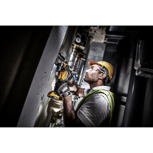 A construction worker wearing a yellow hard hat and safety vest is using a DEWALT cordless oscillating multi-tool to cut or grind into a wall. The worker is holding the tool with both hands and is focused on the task in an industrial setting.