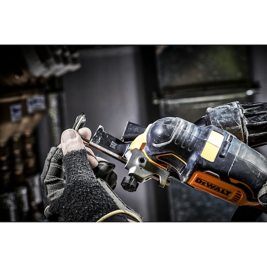 A close-up of a person wearing work gloves using a DEWALT DCS356N oscillating multi-tool for cutting or shaping a metal object in an industrial setting.