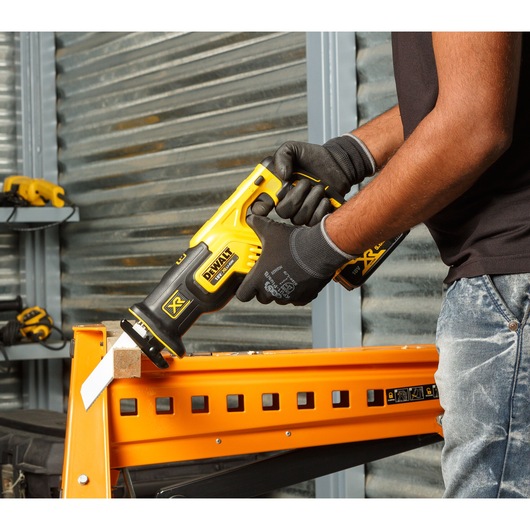 A person wearing black gloves is using a yellow and black DEWALT reciprocating saw to cut wood on an orange metal stand in a workshop.