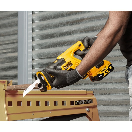 A person wearing gloves is using a yellow and black DEWALT cordless reciprocating saw (model DCS367P2) to cut through a metal pipe placed on a workbench.