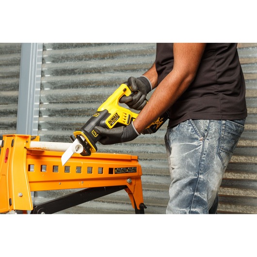A person is using a DEWALT DCS367P2 cordless reciprocating saw to cut through a white pipe held on a yellow workbench. The person is wearing gloves and jeans, and the setting appears to be an industrial or workshop environment.