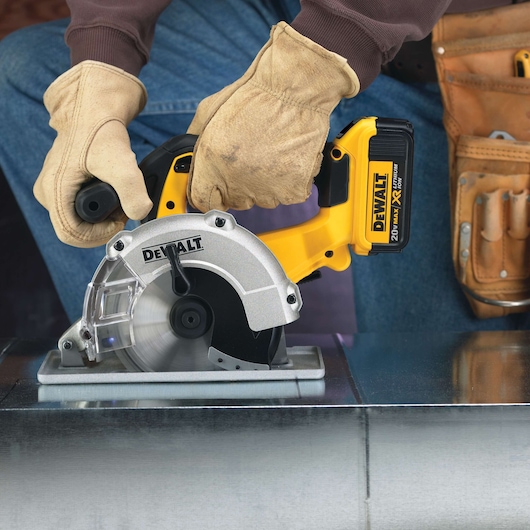 Person wearing work gloves using a DEWALT cordless circular metal-cutting saw on a metal surface. A tool belt is visible on their waist.