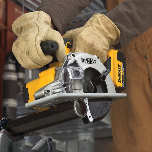 Close-up view of a person wearing work gloves using a DEWALT cordless metal cutting saw to cut through a metal pipe. The tool is yellow and silver with black accents, and the battery pack is visible.