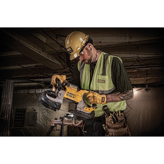 A worker wearing a yellow hard hat, safety vest, and gloves is using a DEWALT DCS374P2 portable band saw on a construction site. The worker's face is blurred for privacy.