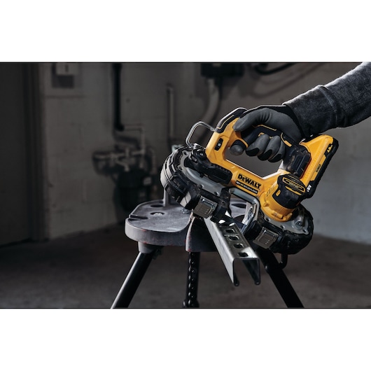 A person wearing gloves is using a yellow DEWALT cordless portable band saw to cut a metal piece that is resting on a workbench in an industrial setting.