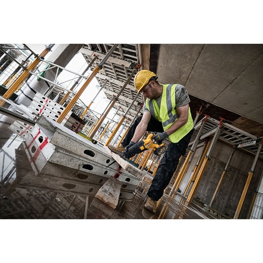 Worker uses DEWALT DCS388N saw to cut wood at a construction site with scaffolding and safety gear.