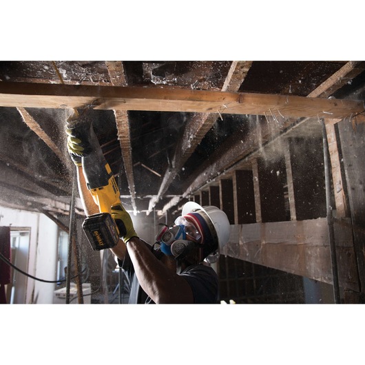 A construction worker wearing safety gear, including a hard hat, goggles, gloves, and a respirator mask, is using a DEWALT cordless reciprocating saw (SKU: DCS388T2) to cut through a wooden beam in a building under renovation.