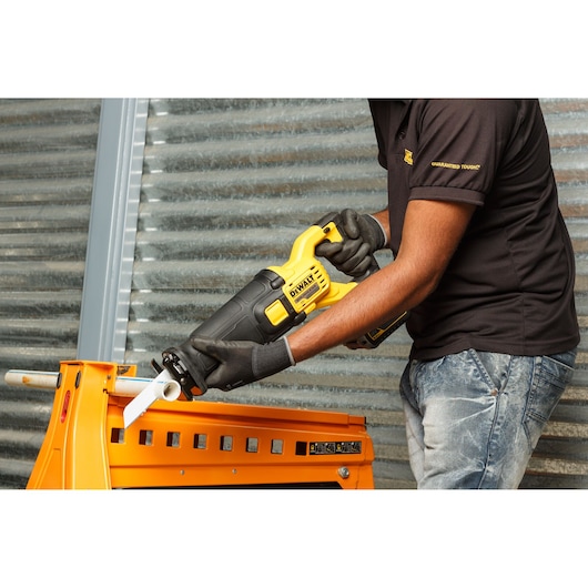 A person wearing work gloves and a black shirt is using a DEWALT cordless reciprocating saw to cut a white pipe, with the saw positioned on an orange workbench in a workshop setting.