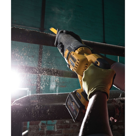 A person wearing gloves uses a DEWALT cordless reciprocating saw to cut through a metal pipe. Sawdust and particles are visible in the air, and there is bright light in the background.